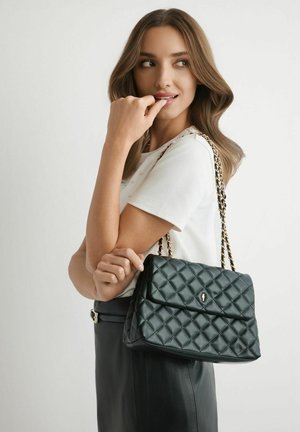 Kobieta o falowanych brązowych włosach, ubranа w białą koszulę i czarną spódnicę, trzymająca czarną pikowaną skórzaną torebkę z złotym łańcuszkiem.