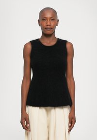 Moteris su nuskusta galva, apsirengusi juoda be rankovių pūkuota palaidine ir laisvomis šviesiai grietinėlės spalvos plisuotomis kelnėmis, stovi prieš paprastą baltą foną.