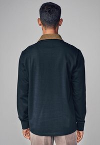 Cămașă închisă la culoare, cu mânecă lungă și guler din fâșie bej, textură netedă și fără modele vizibile. Spatele cămășii are o margine curbată.