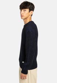 Maglione a costine blu navy con una leggera lucentezza, con scollatura rotonda e maniche lunghe, indossato sopra pantaloni chiari.