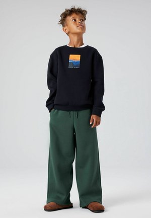 Black sweatshirt with mountain graphic, paired with green wide-leg sweatpants. Material appears soft; shirt has ribbed cuffs and a crew neckline.