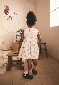 Vestito bianco con stampe colorate di animali e carosello, scollo rotondo e rifinitura rossa. Indossato con calzini rosa e scarpe scure, in piedi su pavimento in legno.