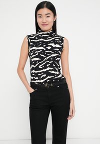 Fekete és krémszínű ujjatlan felső magas gallérral, amely merész, hullámos mintával díszített. Fekete farmerrel és övvel párosítva.