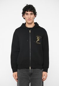 Jaunas vyras su garbanotais plaukais, vilkintis juodą džemperį su auksiniu arklio grafiku ir tekstu, stovi prieš baltą foną.