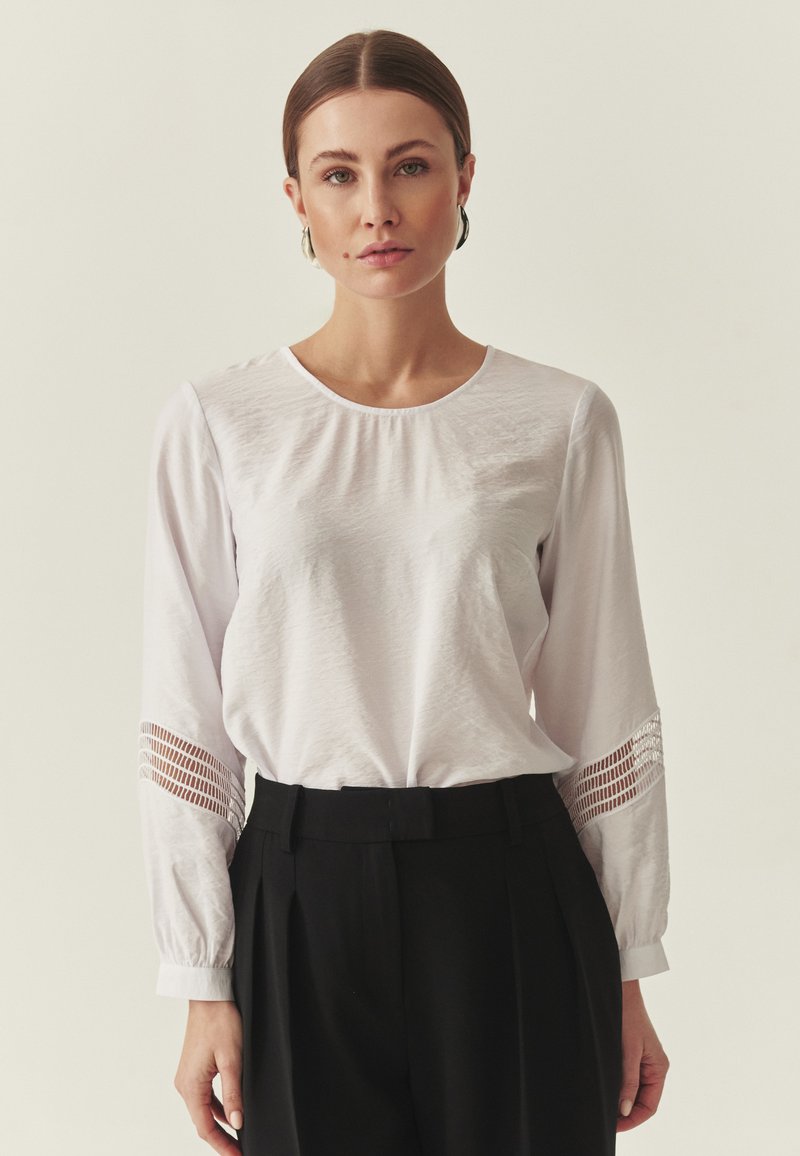 Blouse blanche à manches longues avec un col rond et un tissu texturé. Détails remarquables découpés sur les manches. Associée à un pantalon noir taille haute.