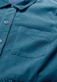 Camicia in corduroy teal con colletto, bottoni frontali e taschino sul petto sinistro. Il tessuto presenta una finitura testurizzata a coste.