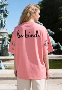 Femme aux cheveux blonds bouclés portant un t-shirt rose oversize avec le texte « be kind. » et des motifs de croix sur les manches, debout près d'une fontaine.