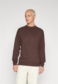 Suéter de punto marrón con manga larga y un cuello alto, con puños y dobladillo acanalados, usado sobre pantalones de color crema.
