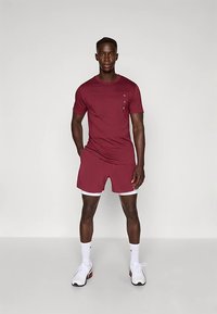 Hombre atlético de pie, vistiendo una camisa de manga corta burdeos y pantalones cortos, calcetines blancos y zapatillas blancas, contra un fondo claro y simple.