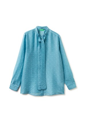 Blouse bleu clair à manches longues avec petits pois blancs, détail noué au col et poignets boutonnés sur fond blanc.