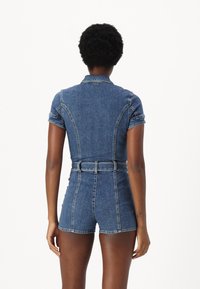 Jeansbyxa med korta ärmar, bälte i midjan och högskurna shorts. Har ljusblått tyg och strukturerade sömmar. Bakifrån visad.