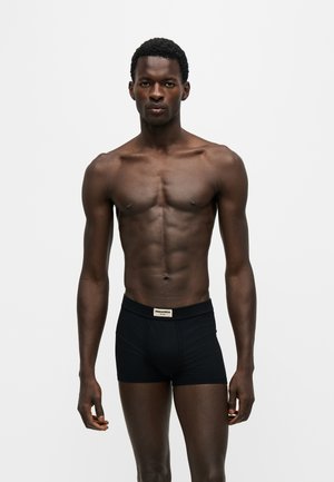 Muskuløs mørkhudet mand iført sorte tætsiddende boxertrusser med et lille hvidt mærke på taljen, stående mod en simpel lys baggrund.