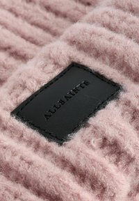 Vaaleanpunainen neulekangas, jossa on teksturoitu, pörröinen pinta, ja musta nahkaleima, johon on kohokuvioitu teksti "ALLSAINTS".