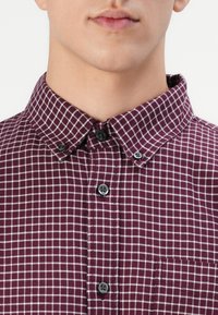 Burgundy checkered shirt made of cotton. Features a button-up collar, front buttons, and a pocket on the left side, with small white checks.