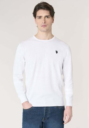 Giovane uomo con capelli scuri che indossa un maglione bianco a maniche lunghe con un piccolo logo scuro sul petto e jeans blu, in piedi davanti a uno sfondo semplice.