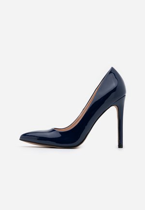 Glinsterende donkeblauwe lakleren pump met hoge hak, puntige neus en slanke naaldhak op effen witte achtergrond.