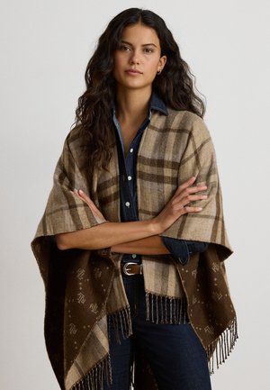 Femme aux longs cheveux foncés porte un châle à carreaux beige par-dessus une chemise en jean foncée et un jean, debout les bras croisés devant un fond uni.