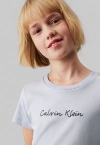 Camiseta de algodón azul claro con cuello redondo y mangas cortas, que presenta un logo en gris oscuro de "Calvin Klein" en la parte frontal.