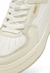 Fehér sneaker, textúrált bőr és velúr részletekkel, lapos fűzőkkel, perforált orral és emelt, párnázott talppal.