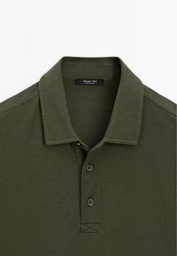 Olivově zelené polo tričko s texturovanou tkaninou, tříknoflíkovou légou a krátkým límcem. Štítek uvádí "Massimo Dutti."