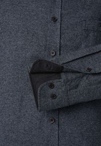 Dunkelgraues Button-Up-Hemd aus texturiertem Stoff mit schwarzen Knöpfen und kontrastierender schwarzer Manschetteninnenausstattung. Der Kragen ist umgeschlagen.