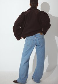 Giacca in suede marrone con maniche ampie, abbinata a jeans a gamba larga blu chiaro con tasche posteriori e un'etichetta di marca visibile. Scarpe marroni.