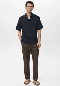 Cămașă cu mânecă scurtă, de culoare bleumarin închis, cu un model texturat, purtată cu pantaloni personalizați maro și sandale maro pe un fundal deschis.