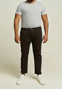 Homme portant une chemise à manches courtes gris clair, un pantalon marron foncé ajusté avec les ourlets retroussés, et des baskets blanches à lacets, debout devant un fond uni.