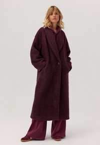 Burgundowy, fakturowany długi płaszcz o oversize'owym kroju, z kołnierzem szalowym, jedno guzikiem i bocznymi kieszeniami; w zestawie z dopasowanymi szerokimi spodniami i butami.