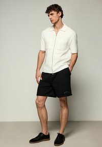 Kortærmet hvid zip-shirt med to frontlommer, parret med sorte shorts. Bæret med sorte sneakers med en tan sål.