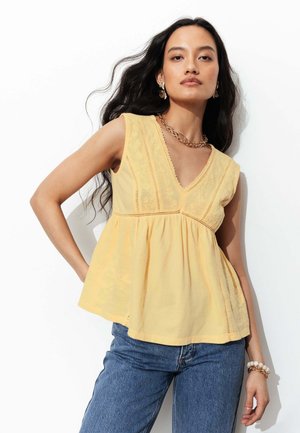 Femme aux longs cheveux foncés portant un haut jaune sans manches à col en V avec broderie et un jean bleu sur fond blanc.