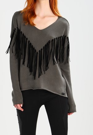 Kobieta w szarym swetrze z dekoltem w kształcie litery V, ozdobionym czarnym frędzlem na klatce piersiowej i rękawach, w połączeniu z czarnymi spodniami.