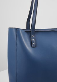 Niebieska syntetyczna torba na ramię o teksturowanym wykończeniu, podwójne mocne uchwyty oraz srebrne nity na paskach. Gładki, zorganizowany design.