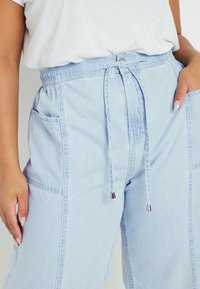 Pantalones de denim azul claro con cintura elástica, cierre de cordón y bolsillos laterales. Presenta costuras visibles y acentos metálicos en las puntas del cordón.