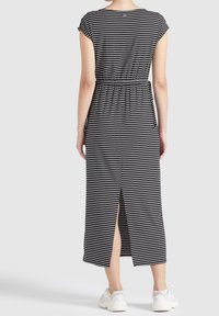 Kvinna i en svart och vit randig midiklänning med sprund bak, kombinerad med vita sneakers, stående mot en vit bakgrund.