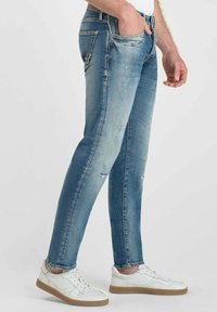 Jeans de mezclilla de corte ajustado en un tono azul claro con un acabado desgastado, detalles rasgados, cinco bolsillos y costuras en contraste. Combinados con zapatillas blancas.