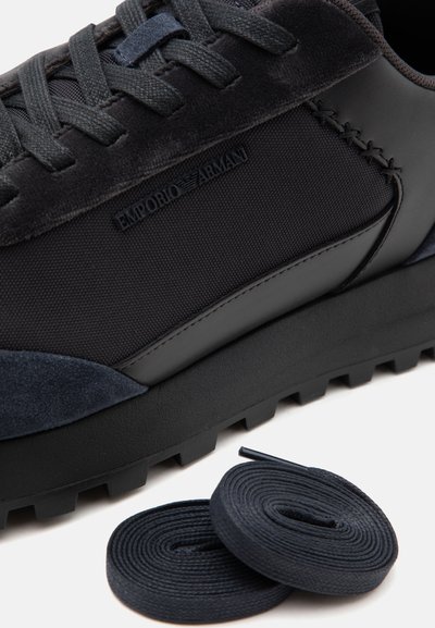 Baskets noires Emporio Armani avec côtés texturés, détails en daim et lacets noirs roulés séparément sur fond blanc.