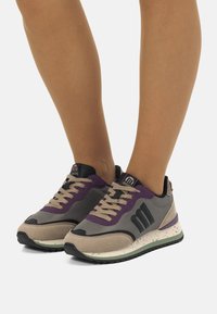 Zapatillas grises y beige con acentos morados, fabricadas con un material texturizado y suela moteada. Presentan un logotipo negro y un diseño clásico de cordones.