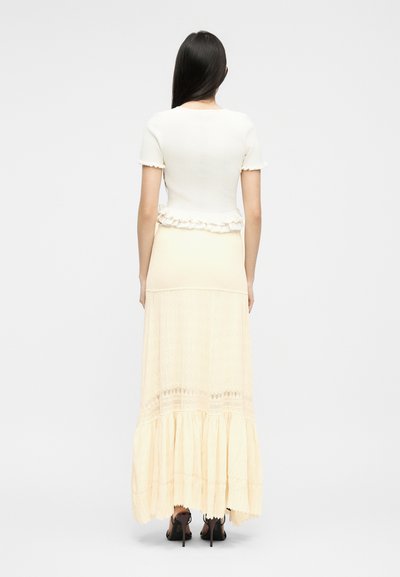 Frau mit langen dunklen Haaren, die ein weißes kurzärmeliges Oberteil mit Rüschenabschluss und einen langen cremefarbenen Rock mit Spitzen-Details trägt, steht mit dem Rücken zur Kamera.