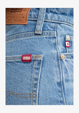 Gai zilu džinsa bikses ar brūnu ādas etiķeti, sarkanu auduma akcentu, zeltītu aparatūru un kontrastējošiem uzšuvumiem gar kabatām.