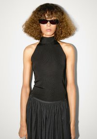 Top nero con scollo all'americana e collo alto, texture a costine. Abbinato a una gonna nera fluida. Gli accessori includono occhiali da sole sottili. Sfondo uniforme.