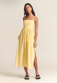 Keltainen maksi-mekko, jossa on neliönmuotoinen pääntie, ohuet hihnat ja vyötärö, joka on rypytetty. Mekossa on sivuhalkiot ja kevyt kangas. Mustat sandaalit.