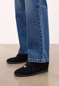 Melnas iešļūcenes ar zelta logo detaļu, kas valkātas ar zilo denimu uz gludas betona grīdas.