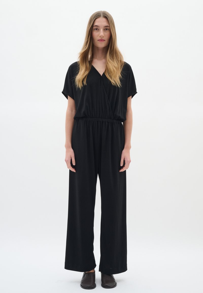 Svart jumpsuit med korta ärmar och V-ringning, med ett elastiskt midjeband och vida ben. Tyget verkar vara mjukt och lätt.