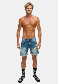 Denimshorts, lyseblå med slidte detaljer, opkrøllede kanter; båret med sorte sokker med hvide hajgrafikker. Lys hud, muskuløs bygning.