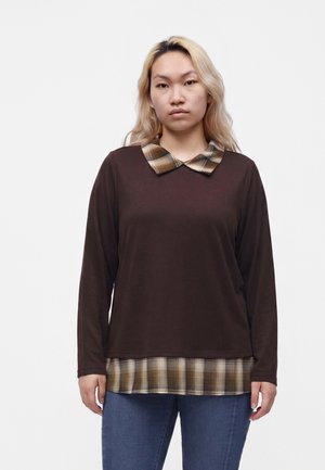 Młoda kobieta z długimi blond włosami, ubrana w brązową bluzkę z długim rękawem, z wzorzystym kołnierzem i brzegiem oraz niebieskie dżinsy, stoi na białym tle.