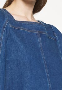 Top in denim con scollo quadrato, colore blu, cuciture a contrasto e vestibilità ampia, con superficie testurizzata e drappeggio sottile.