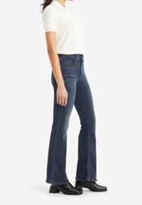 Jean flare bleu marine en denim, taille haute, avec des détails de décoloration subtils, associés à un polo à manches courtes crème et des bottines noires.