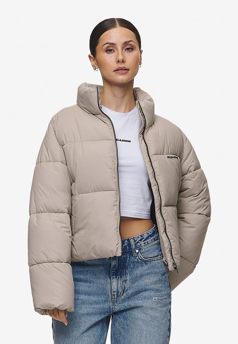 Lys beige puffer-jakke med høy krage, glidelås og løs passform. Har sydd paneler og et logo-detalj på venstre bryst.