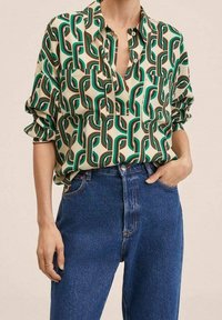 Blouse multicolore en crème, vert et marron avec un motif géométrique, dotée d'un col à revers et de deux poches avant, portée avec un jean bleu.